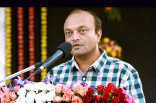 ફૂલોની પાછળ માઇક્રોફોન પર બોલતા મહેશ સવાણી
